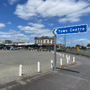 Ngaaruawaahia Community Hub and Library A blue "Town Centre" sign points left above a gray, empty lot with white posts. Behind, buildings line the street, and the sky is bright with fluffy clouds.
