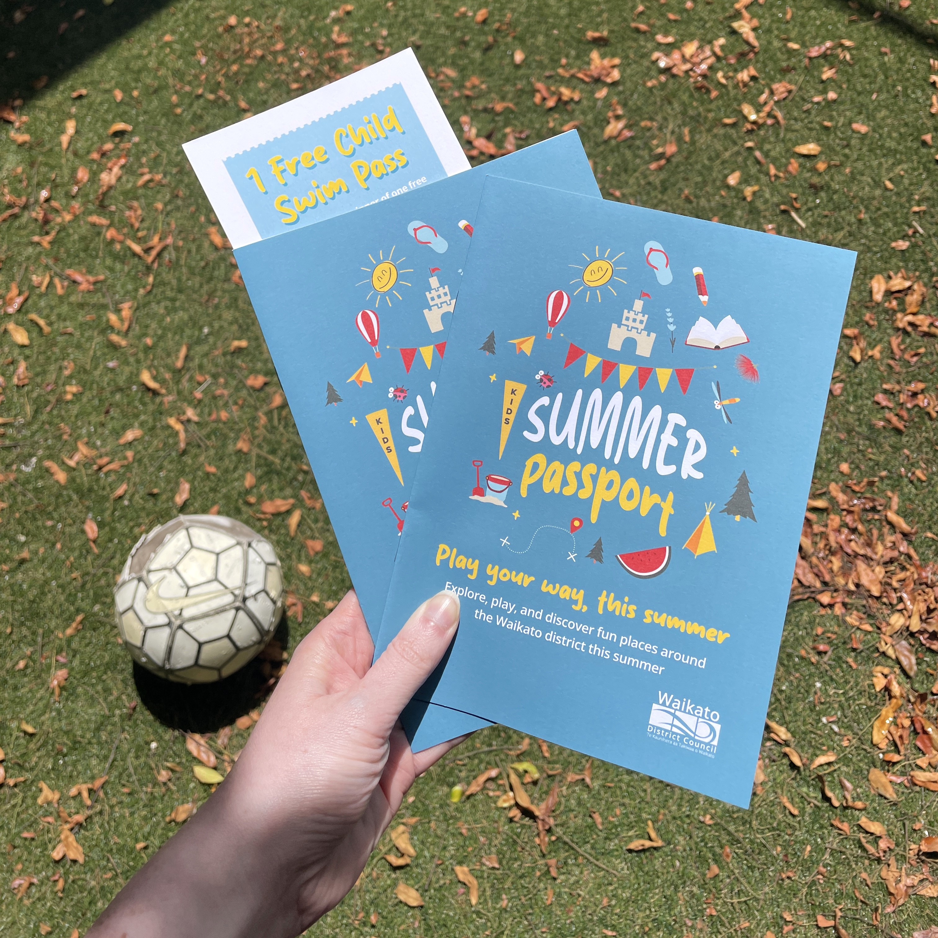 A hand holds two colorful "Summer Passport" flyers with playful graphics and text on a grassy field with fallen leaves and a soccer ball nearby.