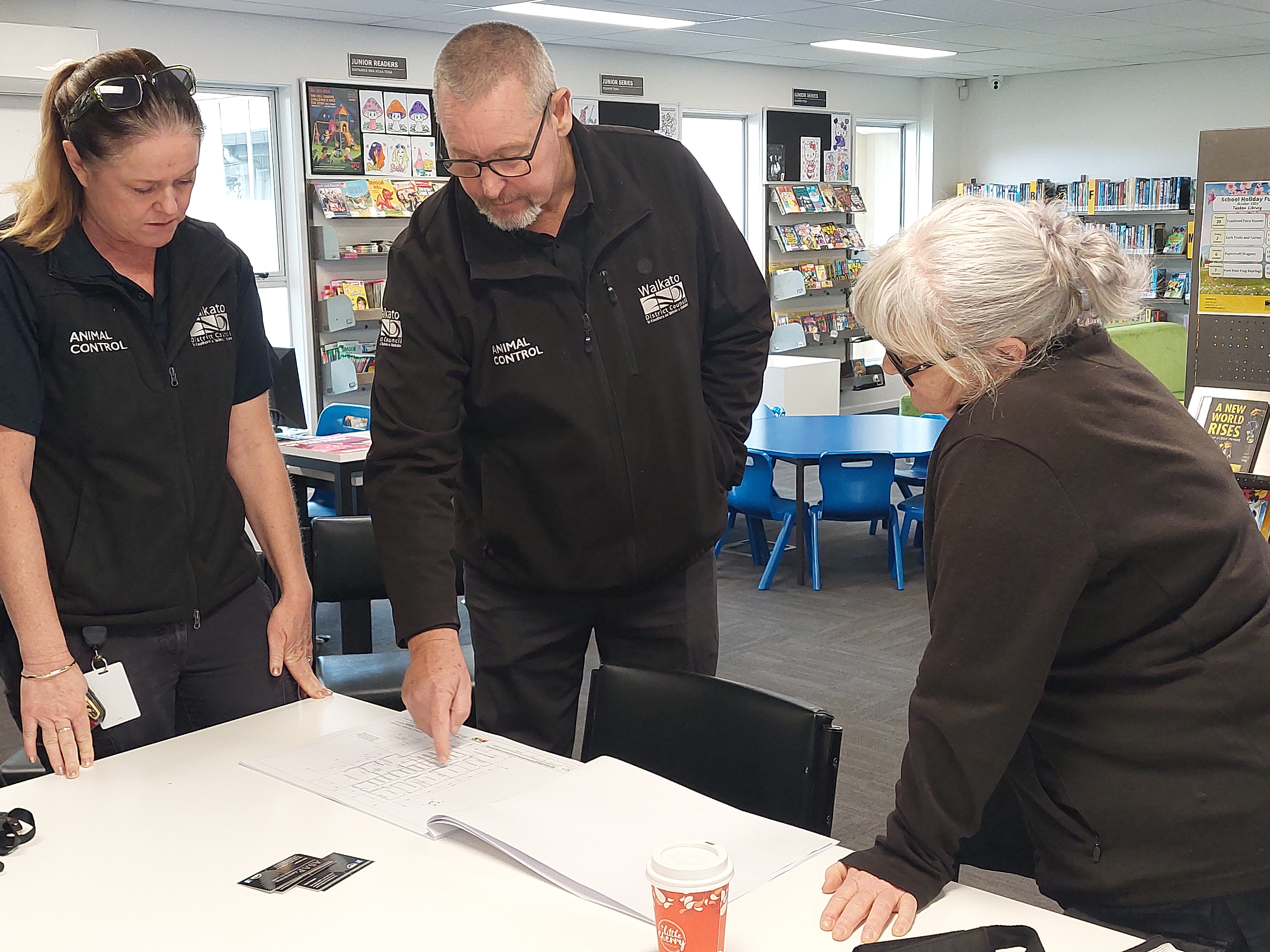 Three staff members in black uniforms lean over a table to review a printed plan in a bright classroom, focused and working together.