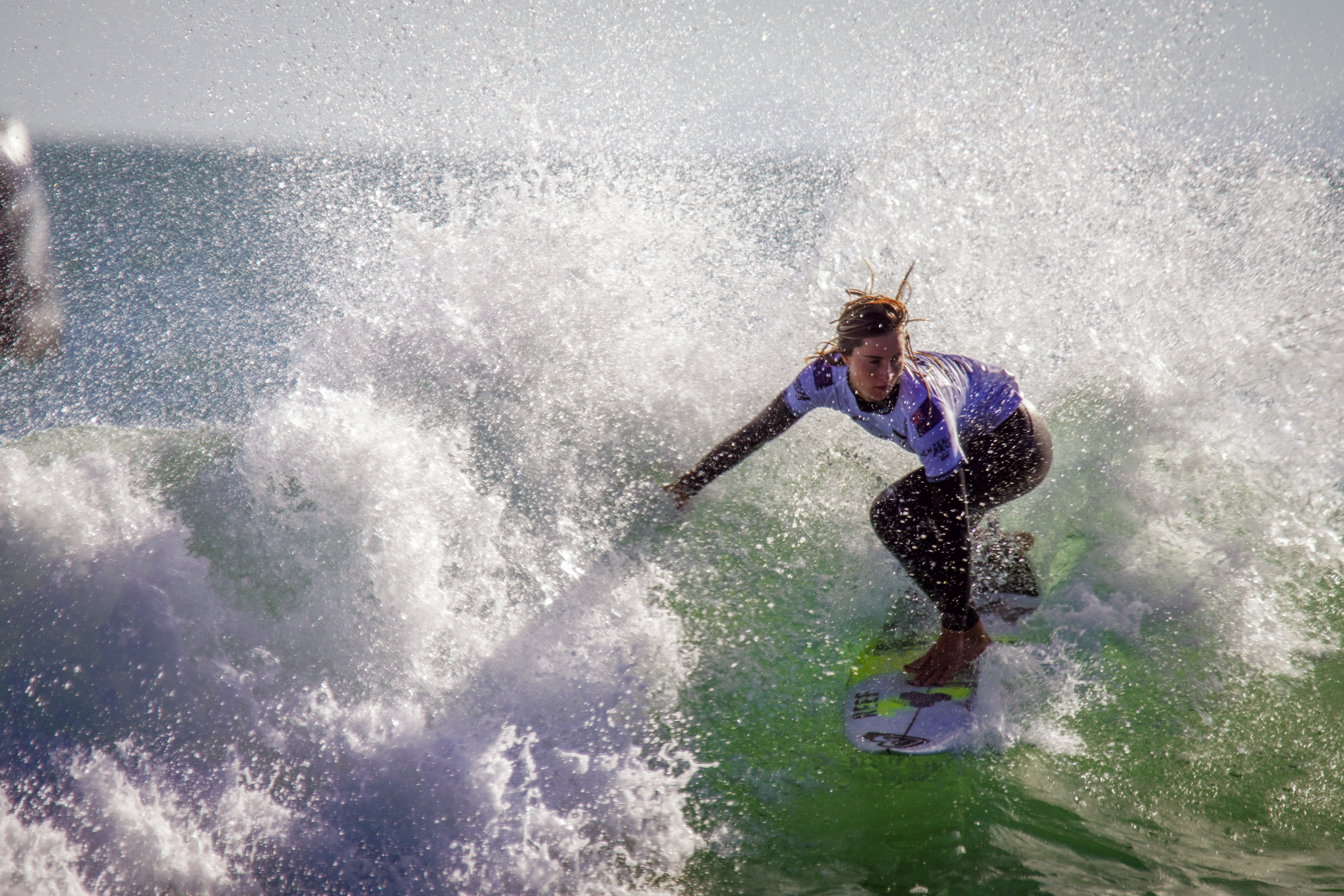 Surfer crouches on a board inside a crashing green wave, surrounded by white spray and sunlight, conveying energy and adventure.