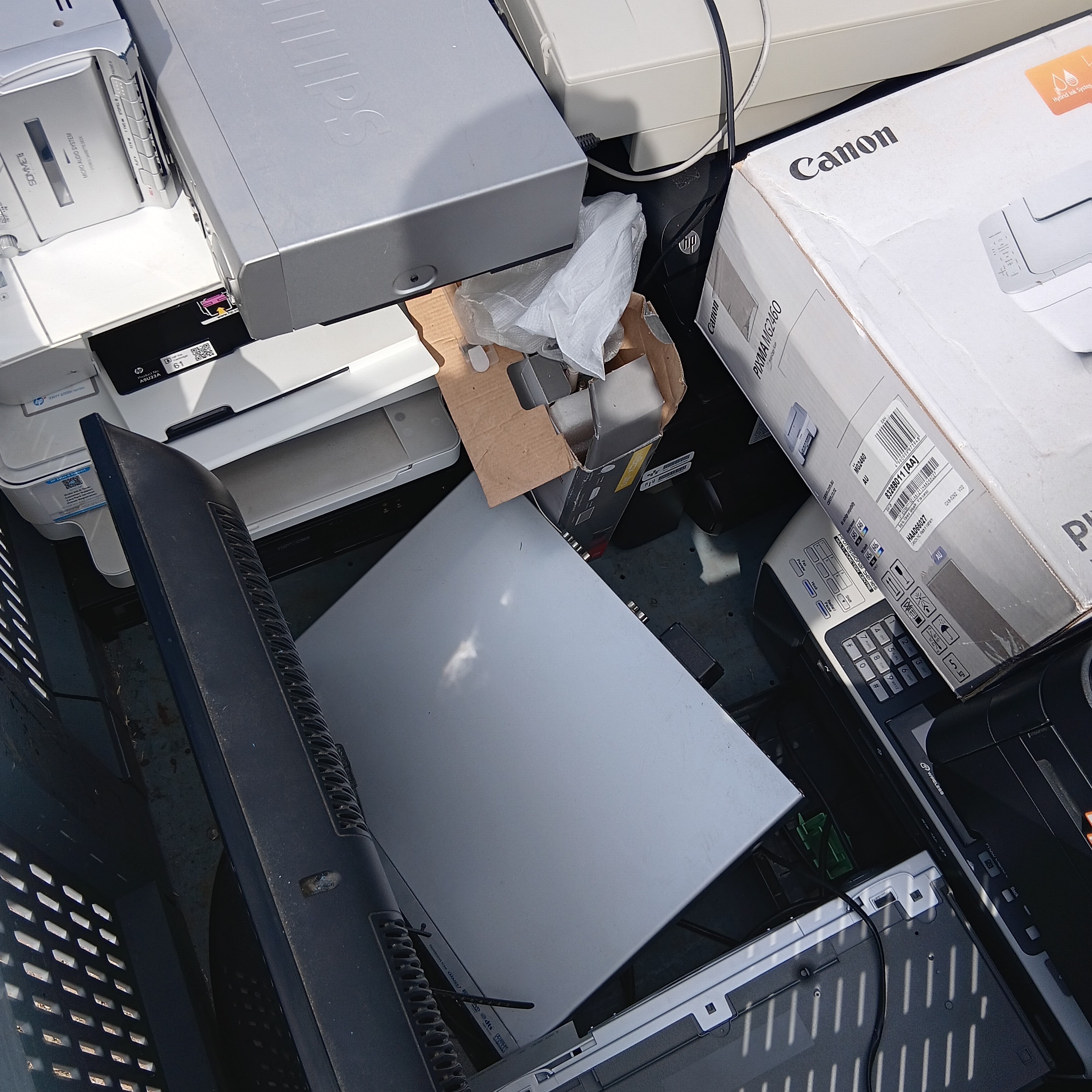 A cluttered collection of discarded printers and monitors, with tangled wires and packaging. The scene conveys disorganization and electronic waste.