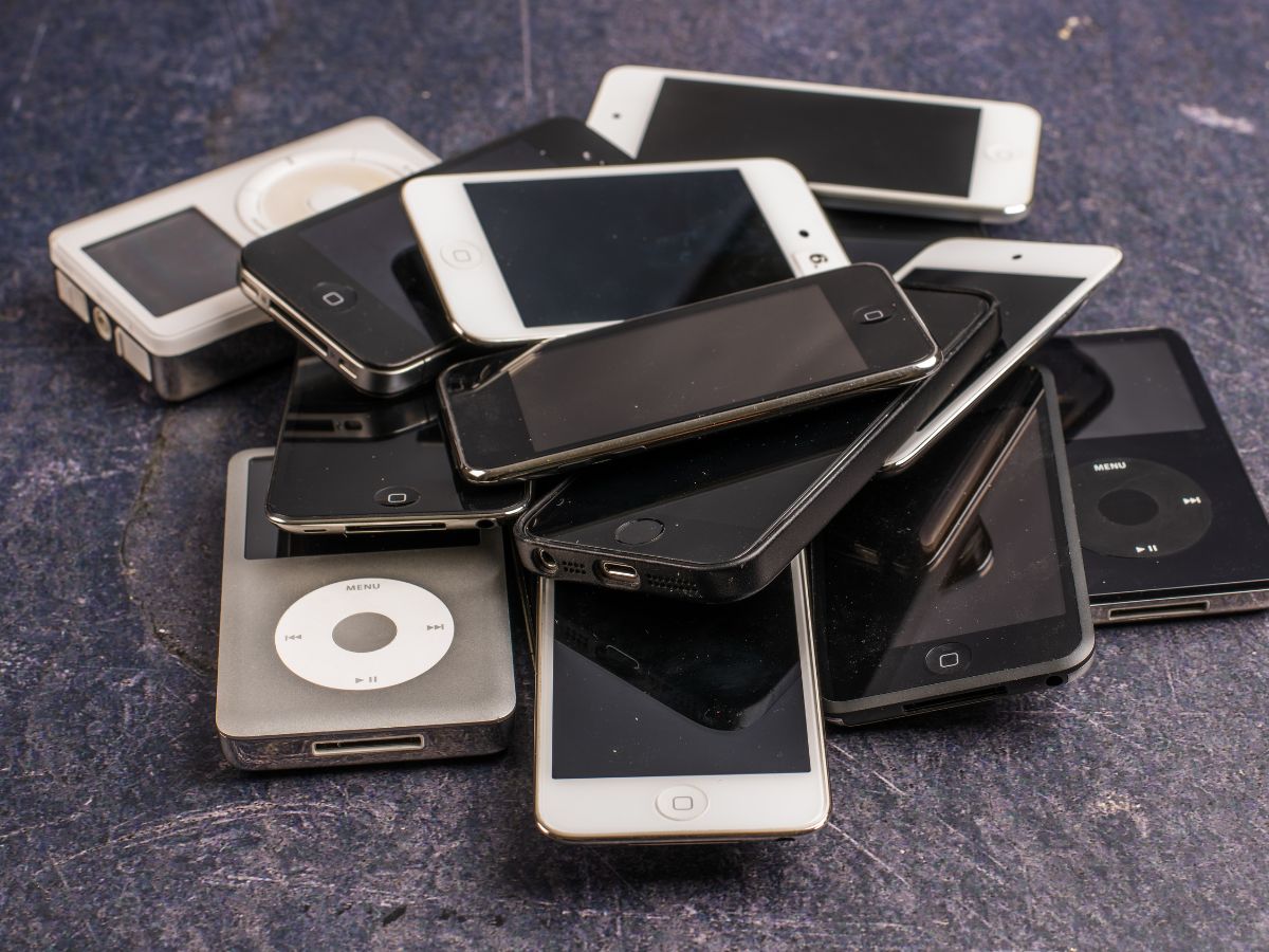 A pile of various black and white iPods and early smartphones on a rough, dark surface. The devices evoke nostalgia for early 2000s technology.