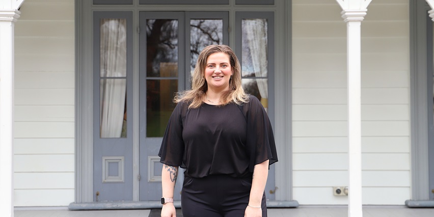 Woman in a black outfit stands on a porch in front of a house with white walls and large windows. She smiles warmly, conveying a friendly tone.
