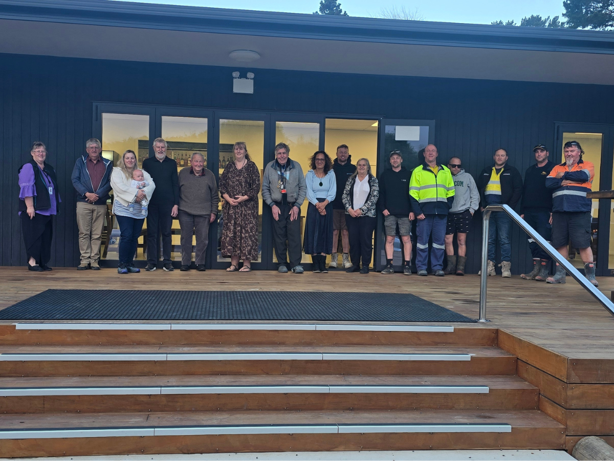 A group of people stands in a line on a wooden deck outside a modern building, smiling. The mood is friendly and casual, under a clear sky.