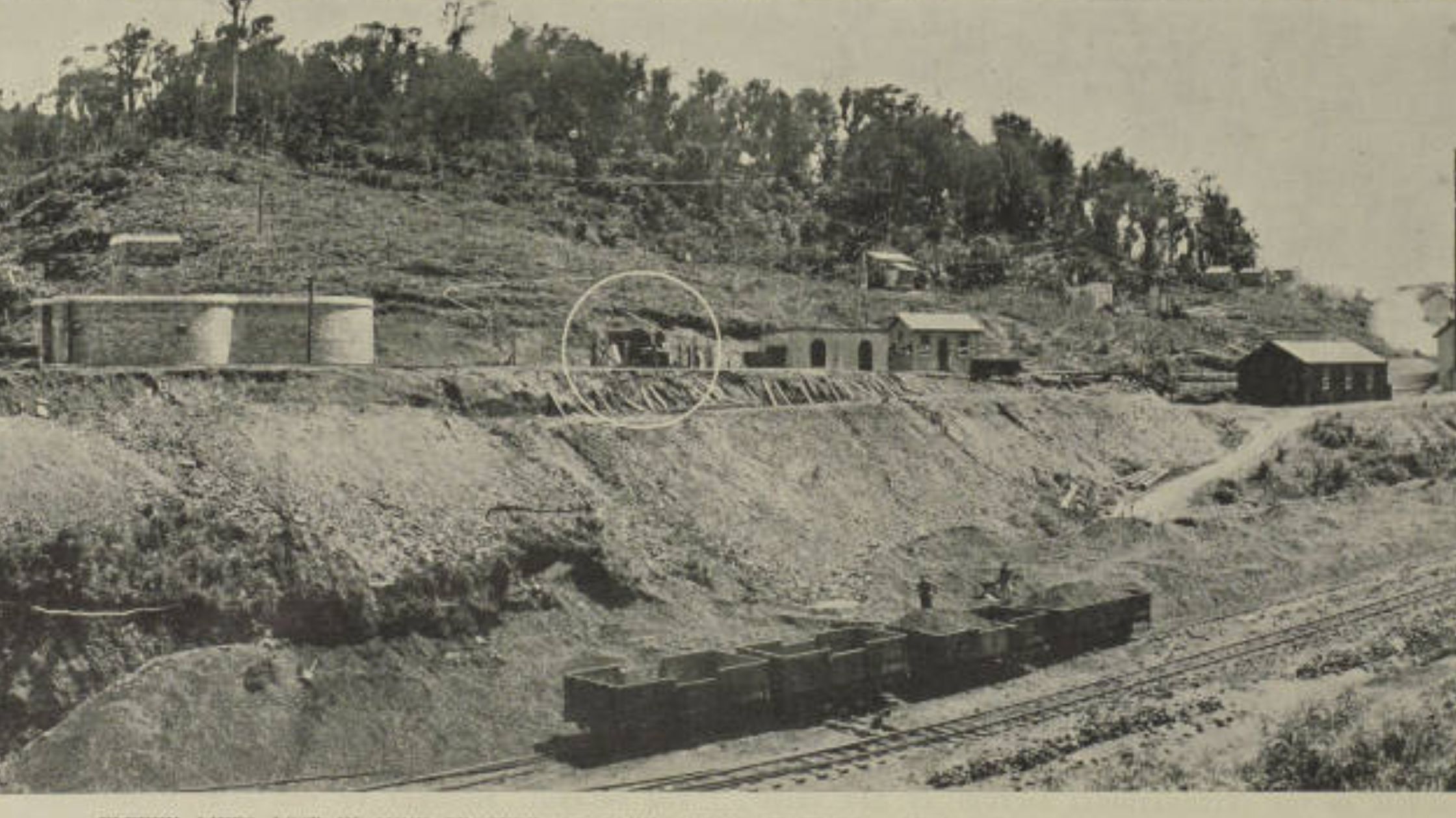 A black-and-white historic photograph shows a hillside with trees, small buildings, and cylindrical tanks. In the foreground, train tracks and several mine carts.