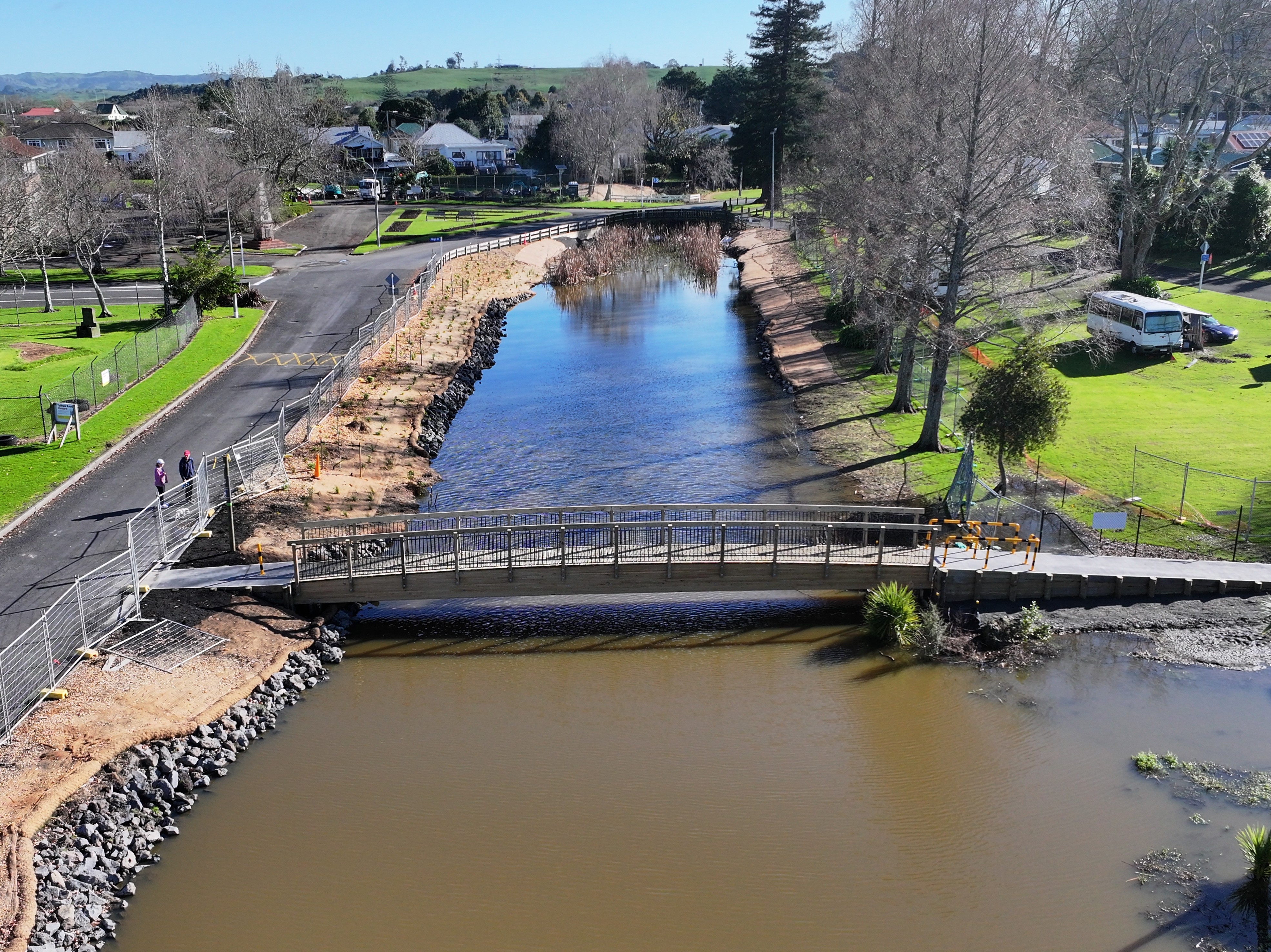 A serene riverside scene with a clear blue sky, featuring a small bridge over a reflective river, lined by trees and a path where two people walk.