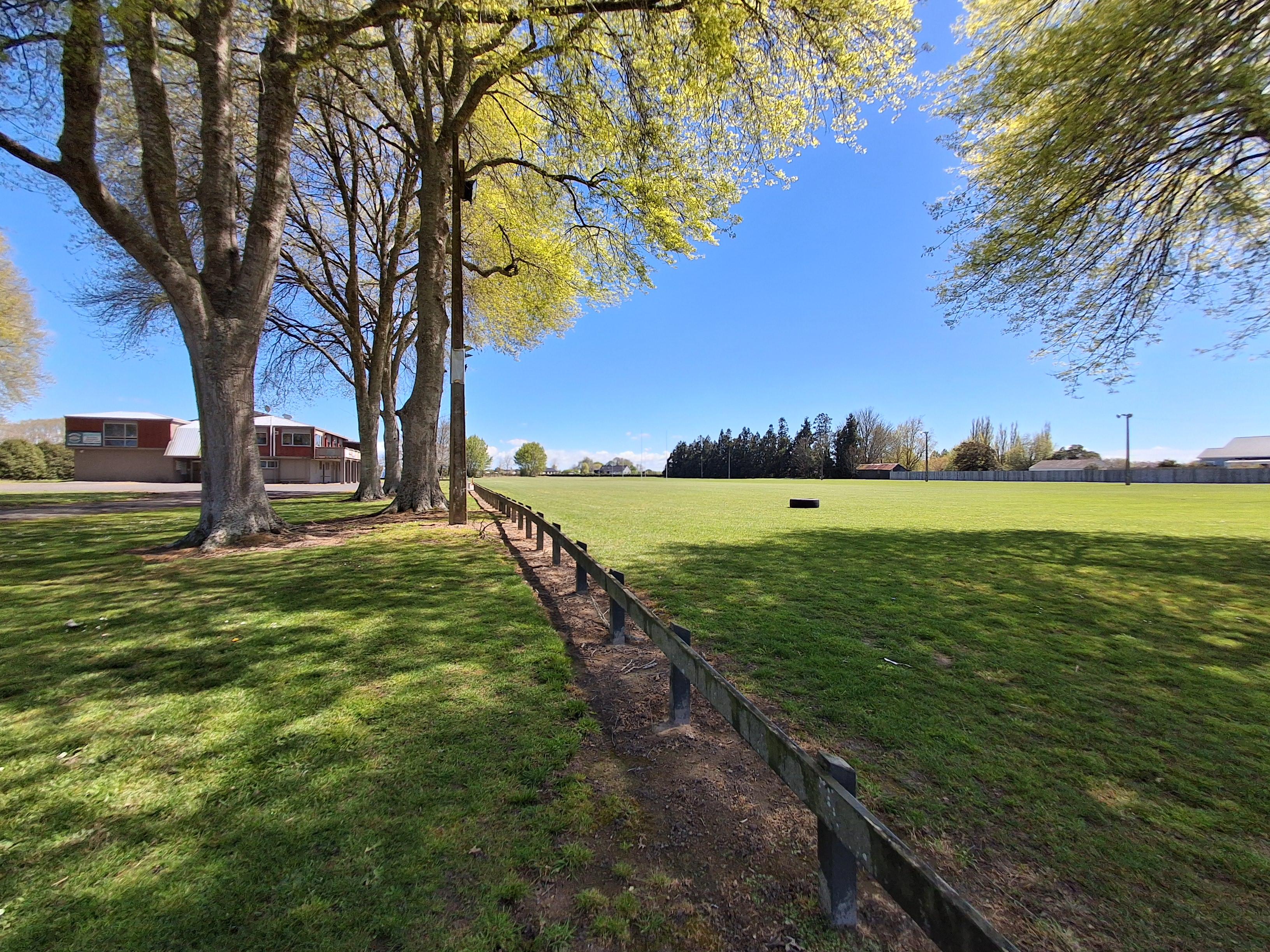 A sunny rural field stretches beneath a clear blue sky, bordered by leafy trees and a wooden fence; a barn and distant woods create a peaceful setting.