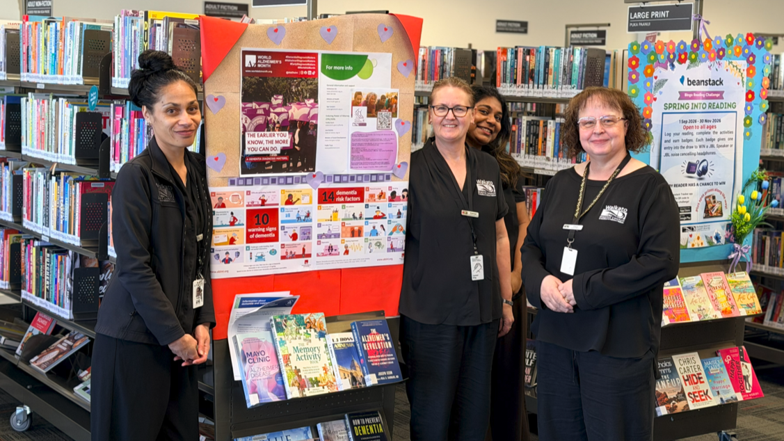 Four library staff members pose beside a colorful community information display and bookshelves, creating a welcoming, cheerful scene.