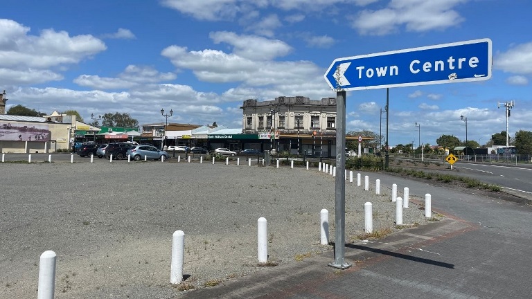 Ngaaruawaahia Community Hub and Library Blue sky with scattered clouds over a town intersection. A sign reads "Town Centre" pointing left. Cars parked around historic building in the background.