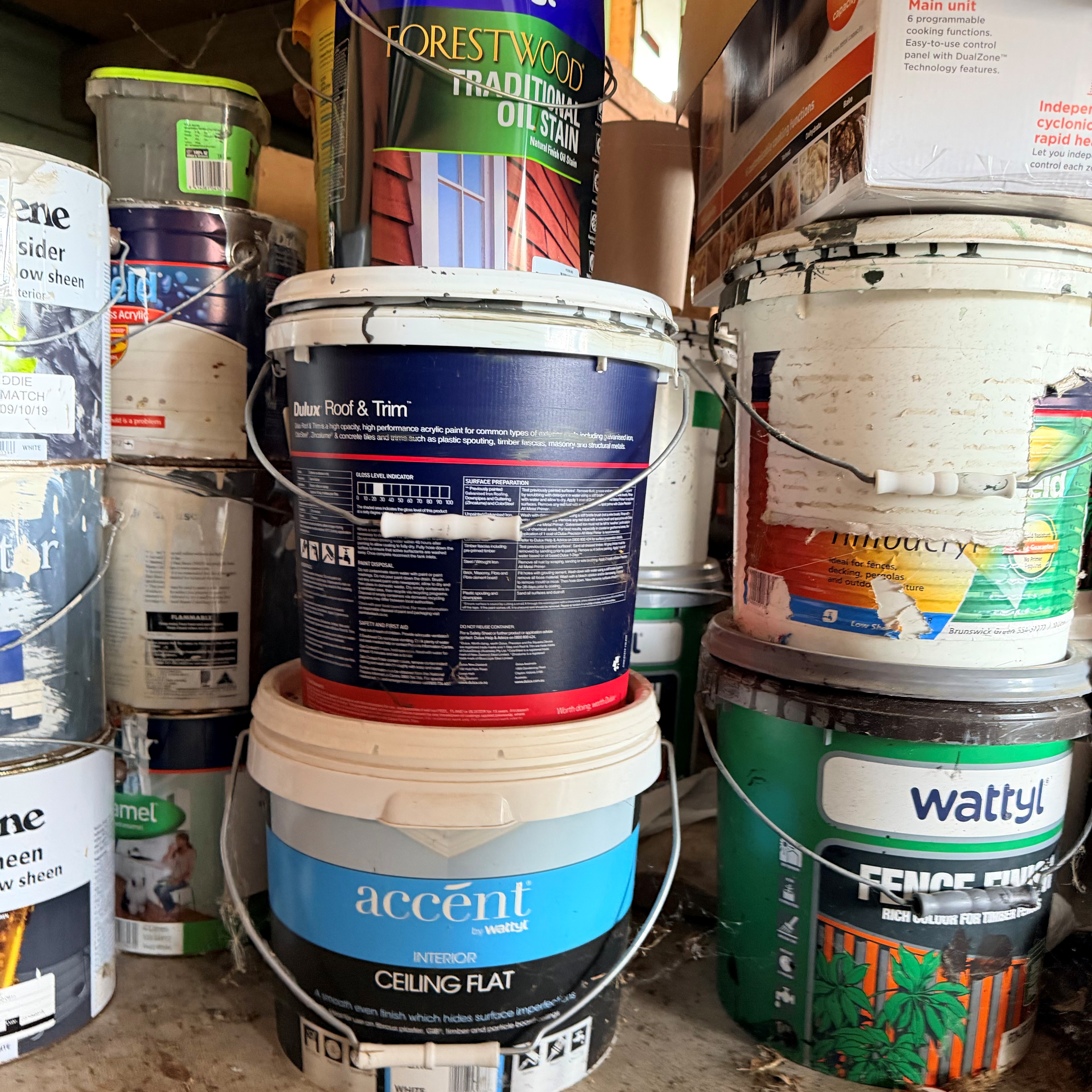 A cramped storage area is packed with stacked paint and finish buckets, including dark blue trim paint and a blue Accent ceiling-flat pail, creating a busy workshop feel.