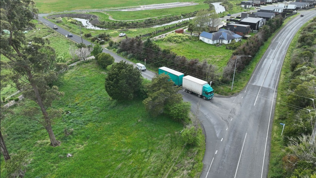 Aerial view of a rural road junction with a turquoise truck, green fields, trees, drainage channels, and scattered homes under cloudy skies.