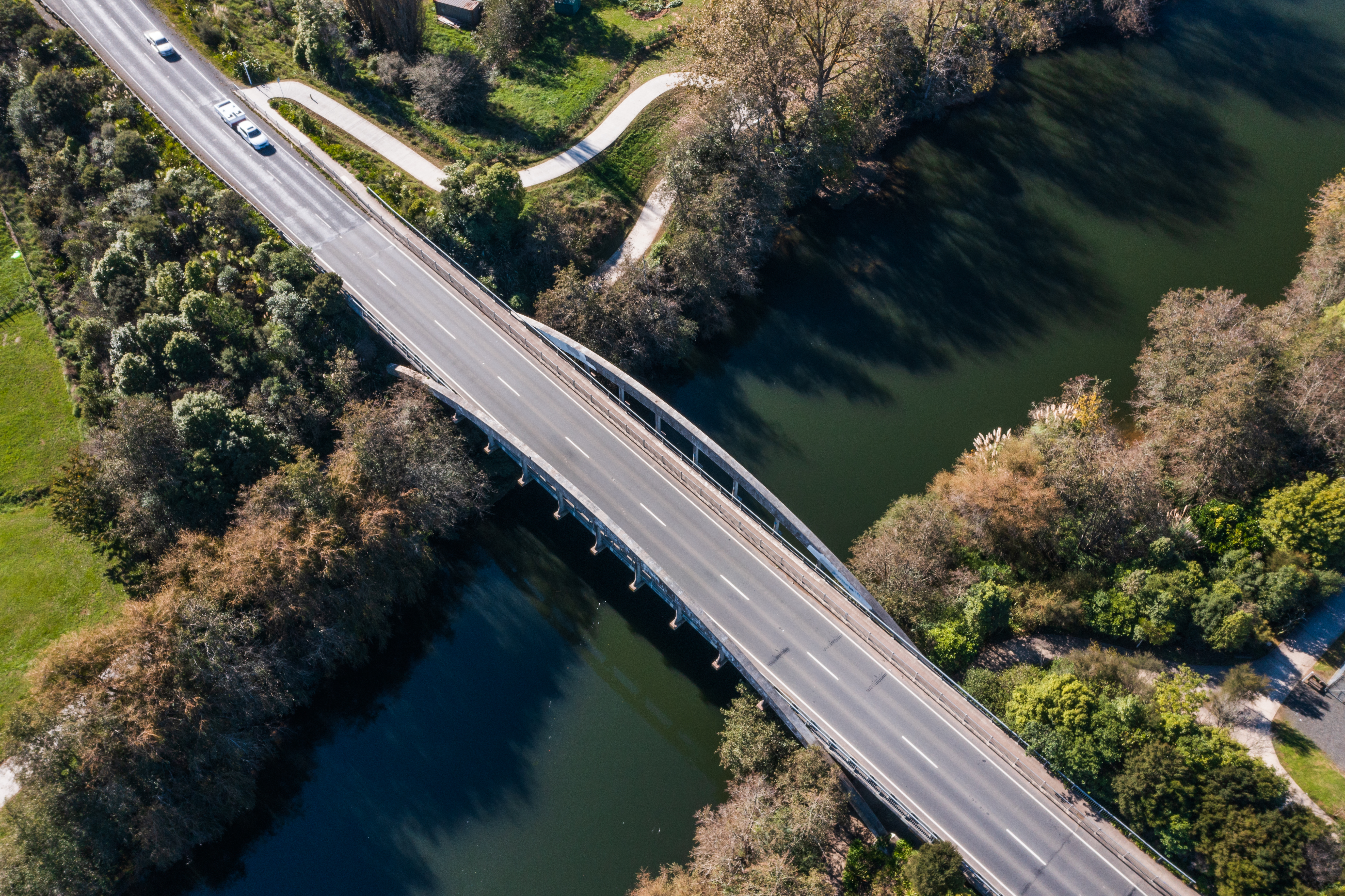 Cars travelling over a bridge that spans over a river, surrounded by greenery and trees, viewed from an aerial perspective.