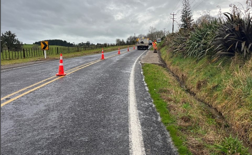 Looking down a sealed, rural road, set up with orange road cones and workers dressed in hi-vis in the distance.
