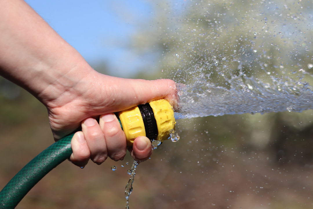Hand tightly grips a green garden hose with a yellow nozzle, spraying water forcefully. Background is blurred, suggesting an outdoor setting.