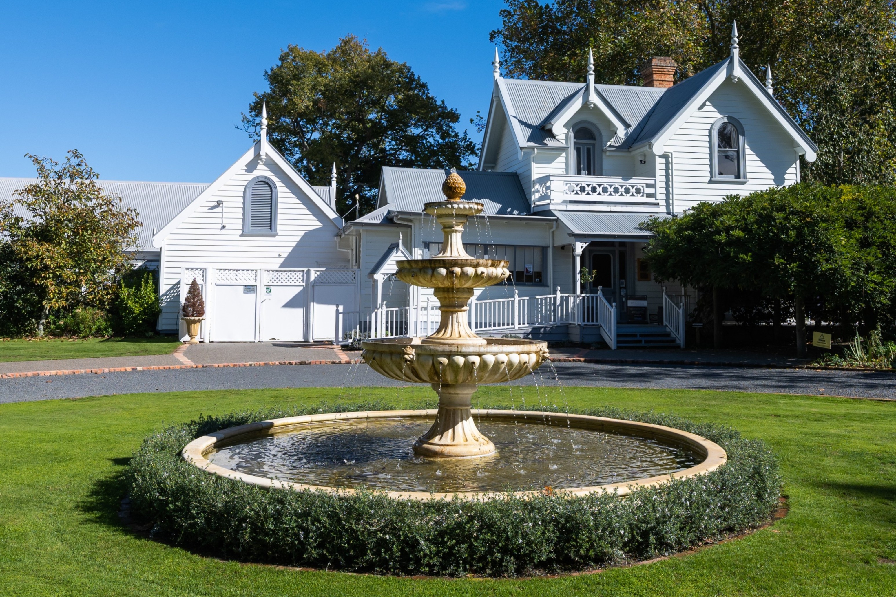 Victorian-style white house with intricate details sits behind a three-tiered fountain. The scene is peaceful, set against a bright blue sky.