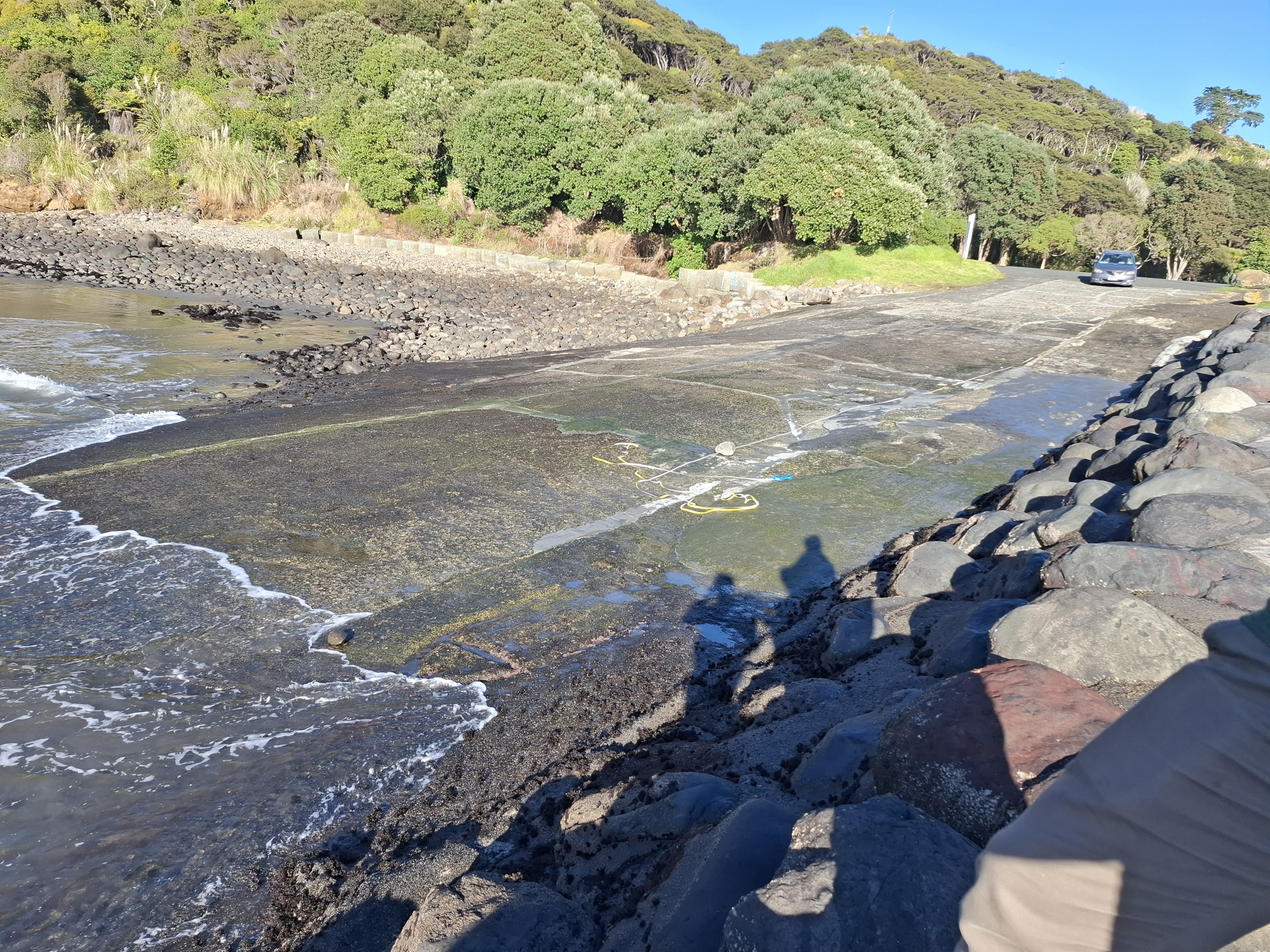 Manu Bay Boat Ramp at low tide, showing the existing concrete ramp extending into the water. Rocky shoreline and coastal vegetation border the ramp, with seawater washing onto the lower section and large rock armouring visible along the edge.