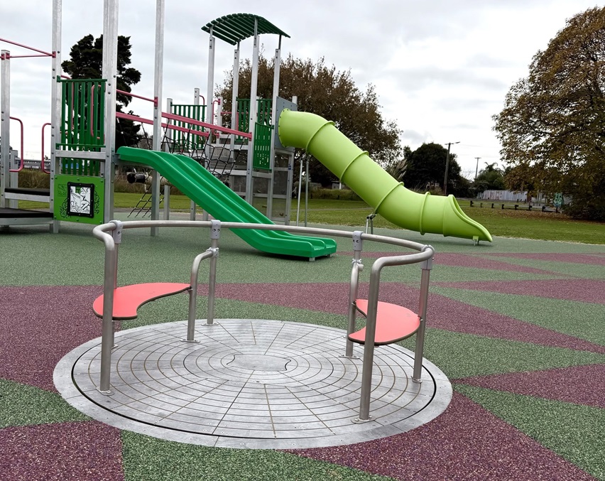 Accessible playground with a ground-level roundabout in the foreground, surrounded by soft-fall safety surfacing.