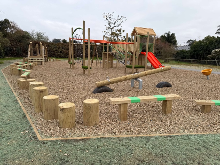 Nature-themed playground with timber balance logs, stepping posts, climbing elements, monkey bars, a seesaw, and a play tower with a bright red slide.
