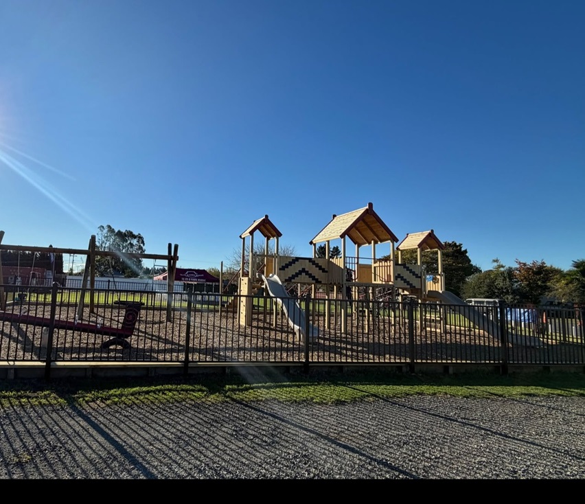 Large fenced playground under a bright blue sky, featuring timber play towers, slides, swings, and climbing structures.