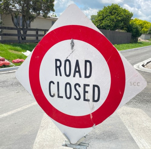 A red and white "Road Closed" sign is positioned on a sunny street. Nearby greenery and a partly cloudy sky convey a calm, daytime setting.