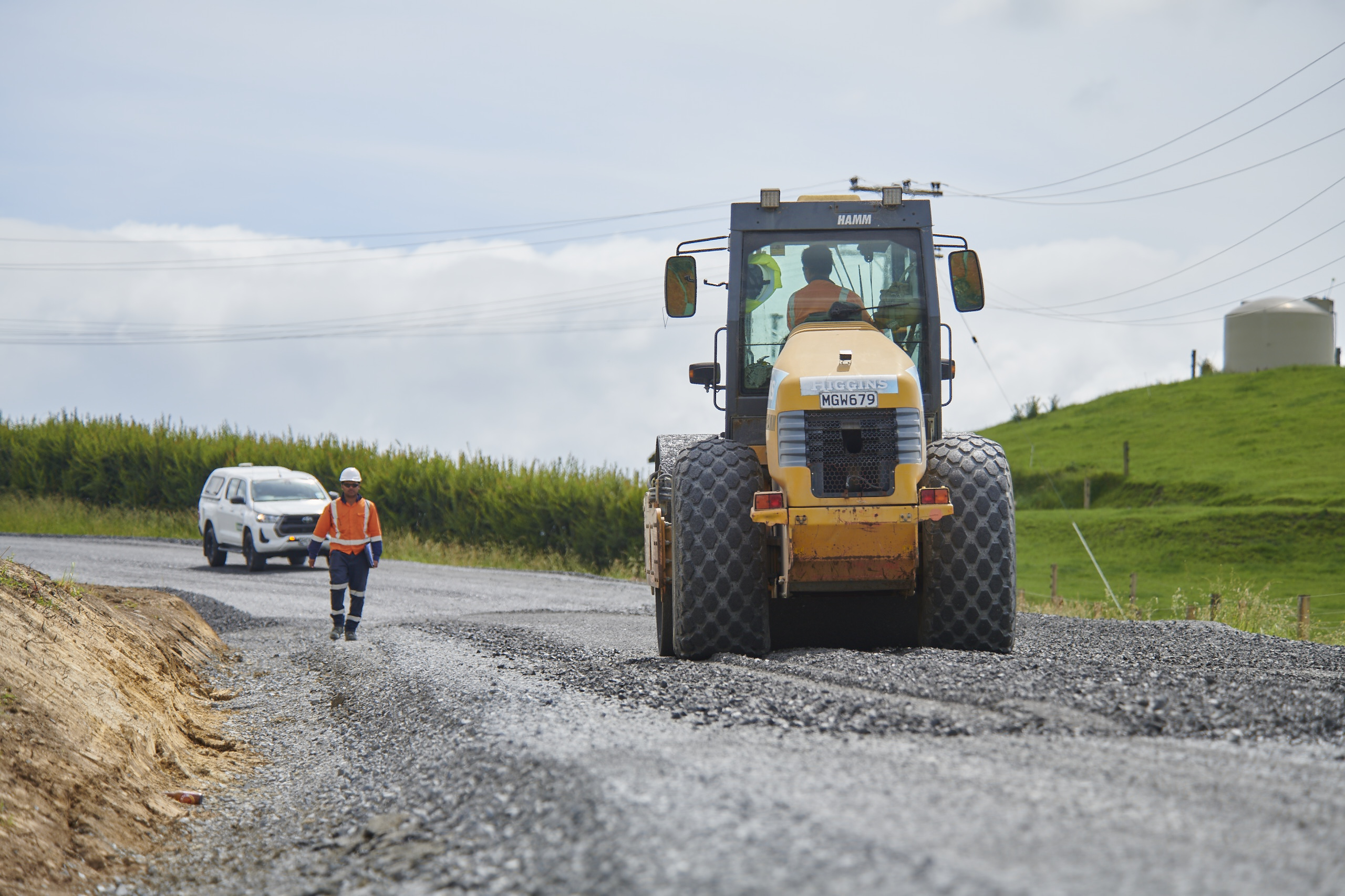 A worker in an orange vest walks beside a yellow road roller on a gravel road. A white vehicle approaches; green hills and a cloudy sky form the backdrop.